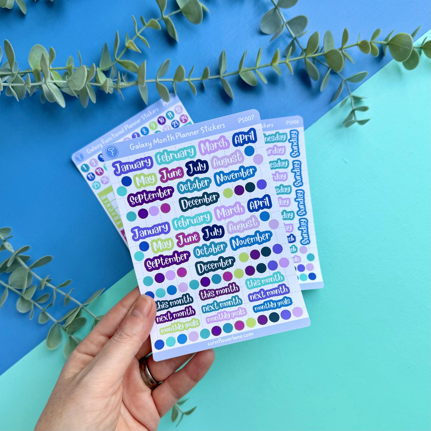 Sheet of planner stickers with months and days text held by a hand on a blue and green gradient background with eucalyptus leaves.