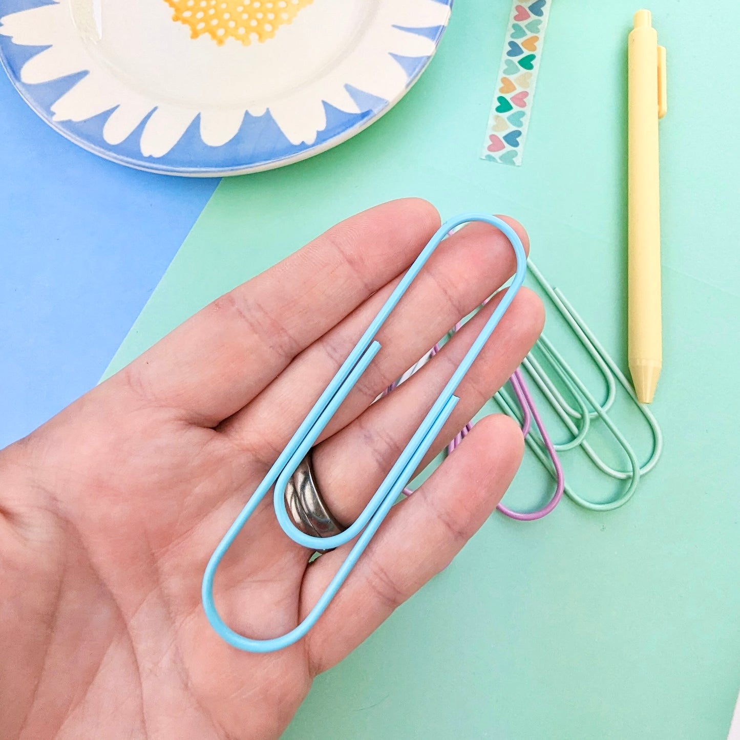Hand holding a blue paperclip with colorful paperclips and a plate in the background